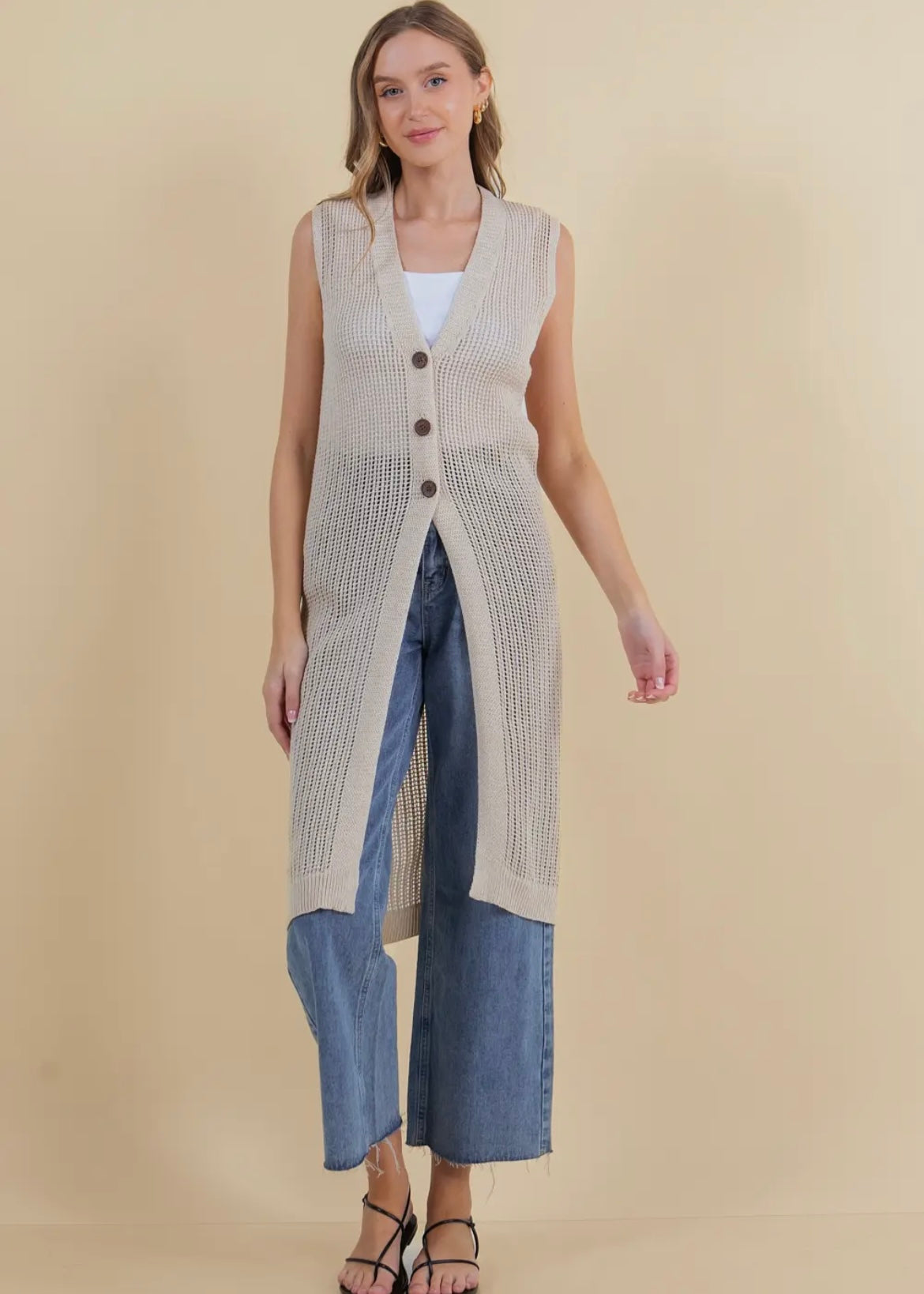 Model showing front view of long open knit sleeveless cardigan in a neutral tan. Cardigan has three button closure.