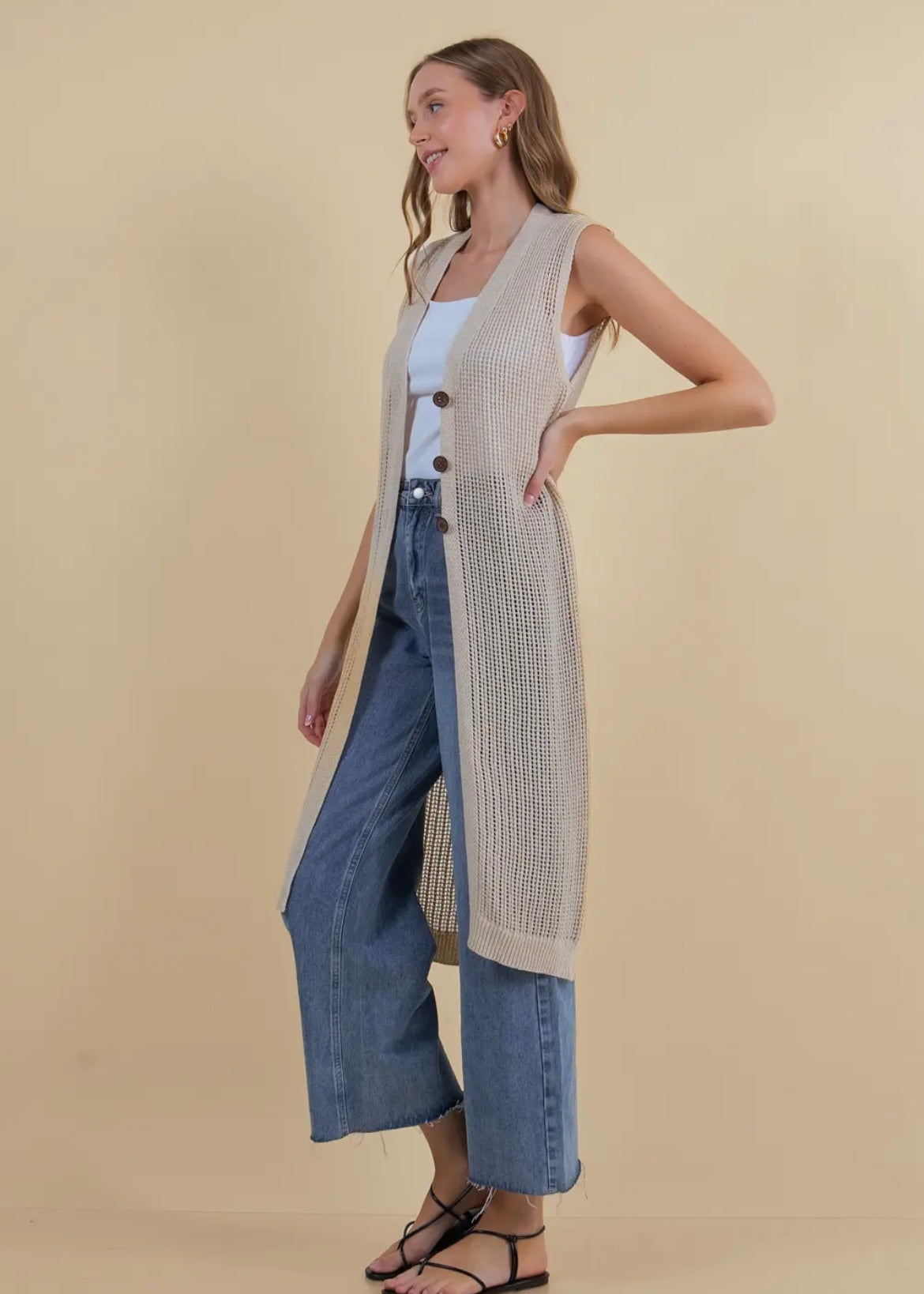 Model showing side view of long open knit sleeveless cardigan in a neutral tan. Cardigan has three button closure.