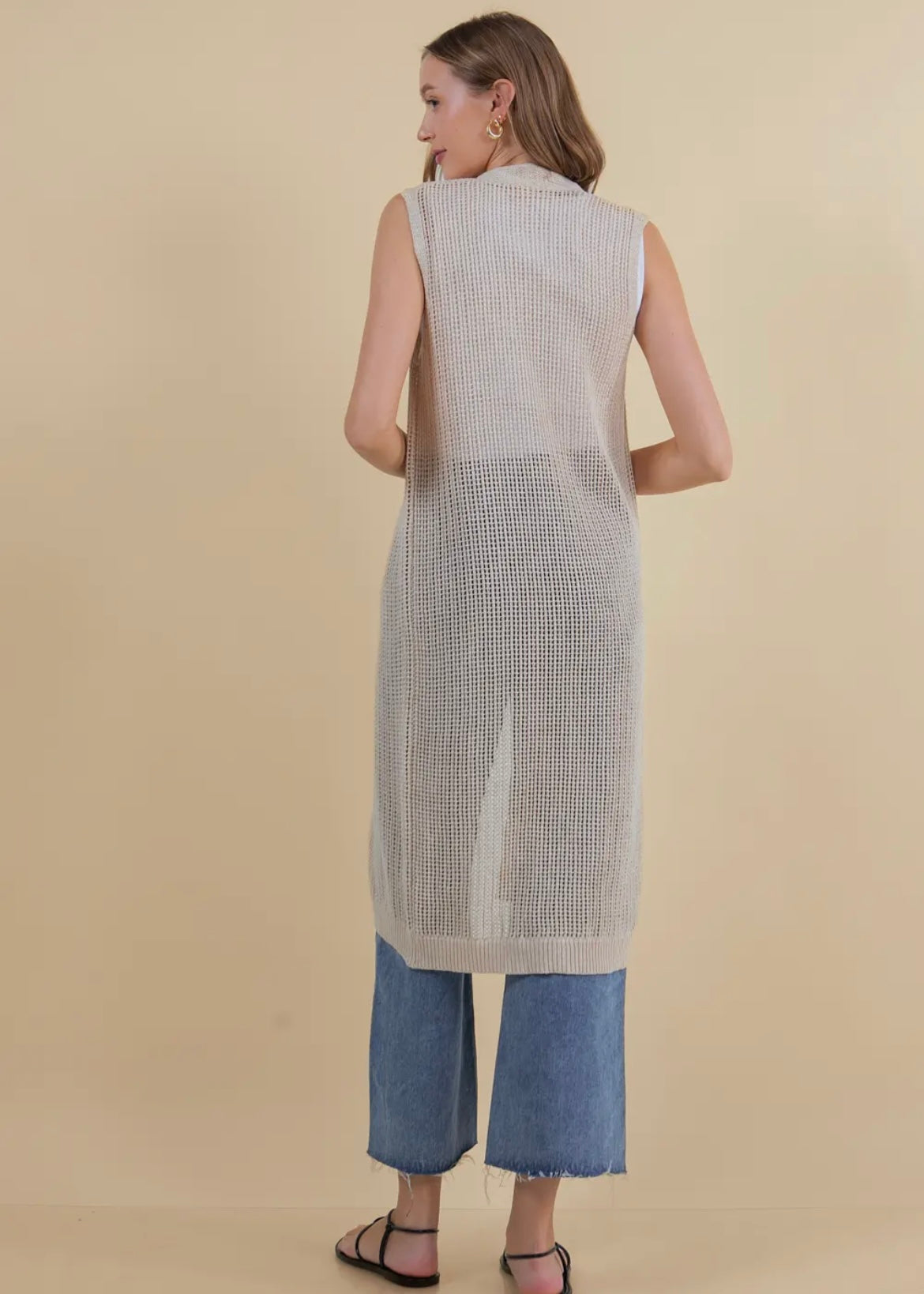 Model showing back view of long open knit sleeveless cardigan in a neutral tan. Cardigan has three button closure.
