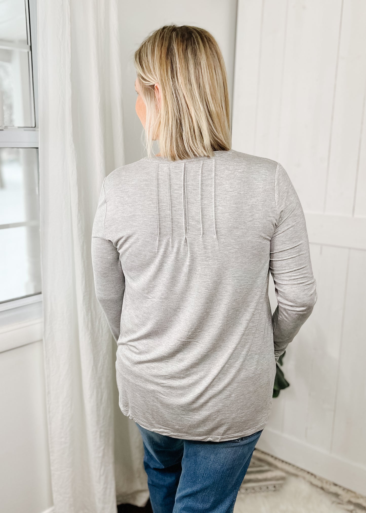 Gray henley top with long sleeves, rounded neck line that goes into a v neckline and a small snap functional button. Top features pin tuck details on both sides for the v neck and o the back in the middle top of the top. 